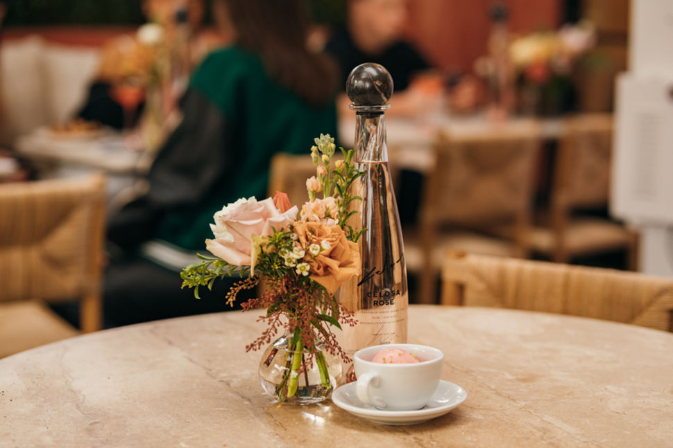 A bottle of Celosa Rosé Tequila with no additives, placed on a table next to a small floral arrangement and a cup of sorbet in a cozy restaurant setting.